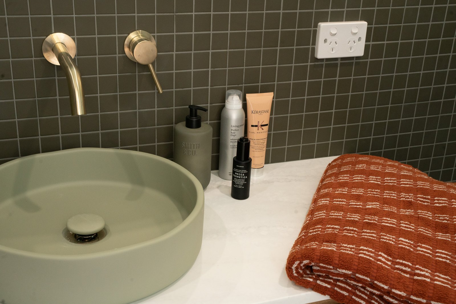 A sage green stone basin under a brushed brass wall tap, dark square tiles and a folded rust towel